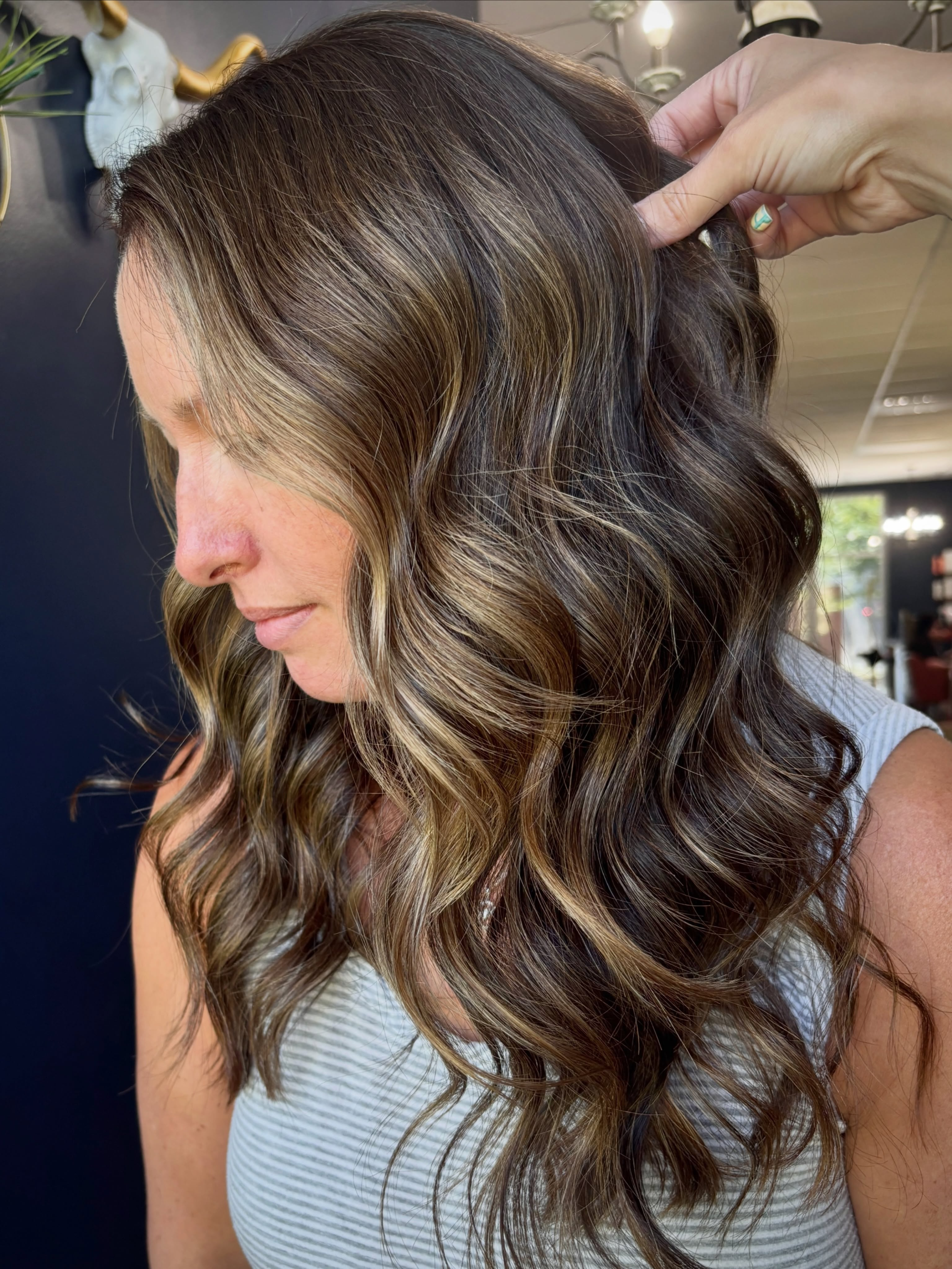 Stylist lifting warm brunette waves with soft caramel highlights at Pearls & Whiskey Salon.
